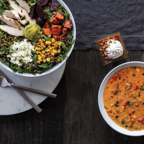 A vibrant salad with chicken, roasted vegetables, and a bowl of soup, accompanied by a dessert in a small dish.