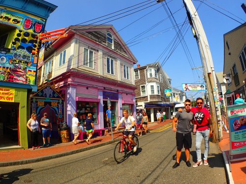Two people pose for a photo on a colorful street with shops, while a cyclist rides by on a sunny day.