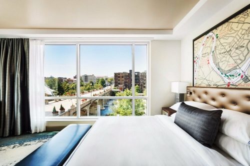 A modern hotel room with a large window showcasing a city view and a comfortable bed with decorative pillows.