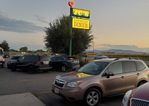 Sign for JC's Country Diner at dusk, with parked cars in the foreground and a scenic landscape in the background.