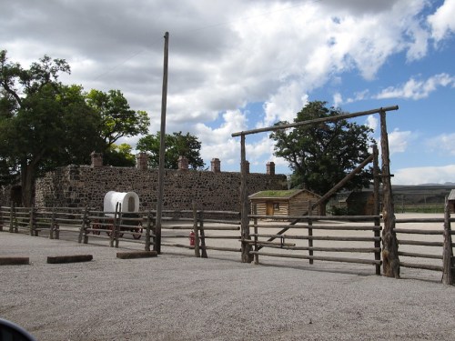 A rustic wooden fence surrounds a historic stone building with a wagon and trees under a partly cloudy sky.