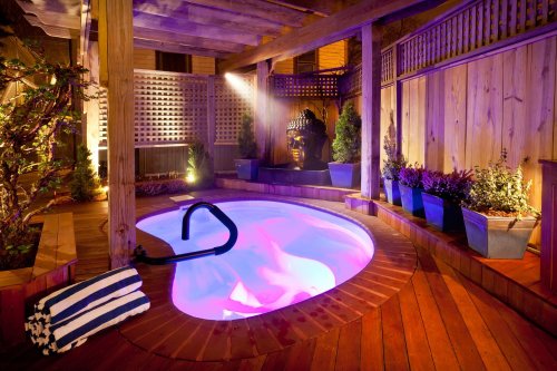 A serene outdoor spa area with a lit hot tub, wooden deck, and decorative plants, featuring a Buddha statue in the background.