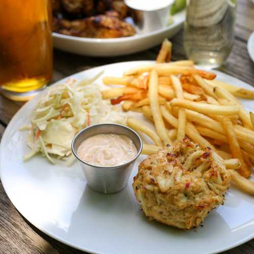 A plate with fries, coleslaw, a crab cake, and a small cup of dipping sauce, with a drink in the background.