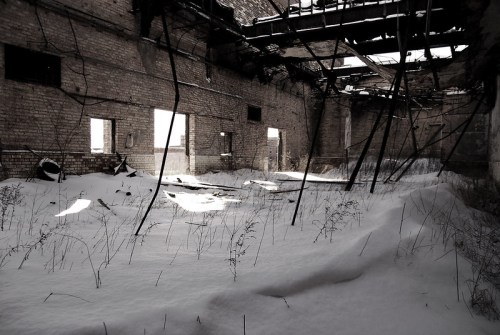 An abandoned building with snow covering the floor and broken structures, illuminated by light through the windows.
