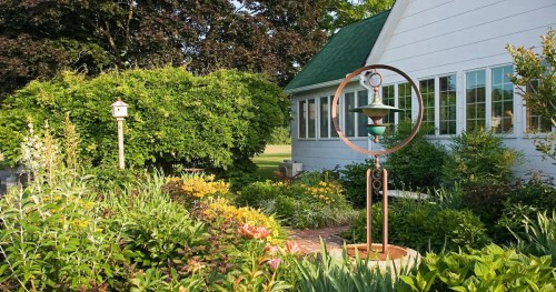 A lush garden with colorful flowers, a birdhouse, and a decorative fountain near a white house with green accents.
