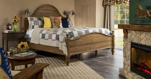 Cozy bedroom featuring a wooden bed, plaid bedding, a stone fireplace, and a sunny window with decorative curtains.