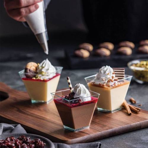 Three elegant dessert cups with whipped cream, chocolate, and fruit toppings, on a wooden board with cookies in the background.