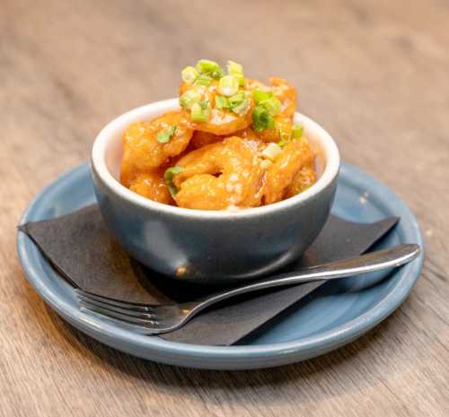 A small bowl of orange shrimp topped with green onions, served on a blue plate with a black napkin and a fork.