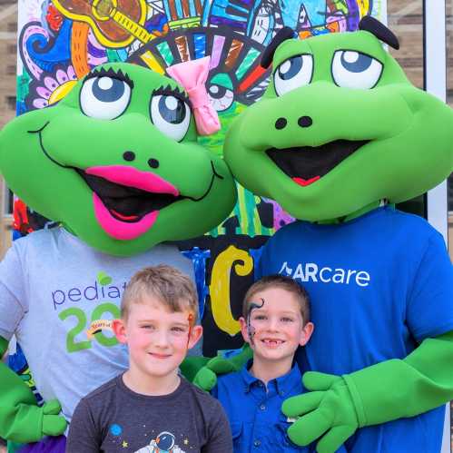 Two costumed frog mascots pose with two young boys in front of a colorful backdrop at a community event.