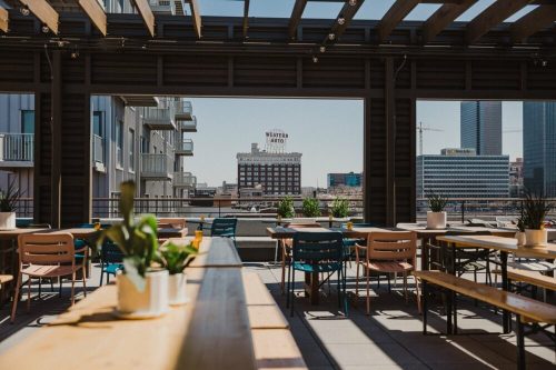 A rooftop dining area with wooden tables and chairs, overlooking a cityscape with a vintage hotel sign in the background.