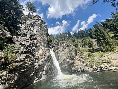 A waterfall cascades down rocky cliffs into a serene pool, surrounded by lush greenery and a blue sky with clouds.