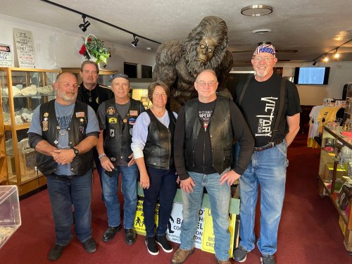A group of six people in leather vests pose in front of a large Bigfoot statue inside a shop.