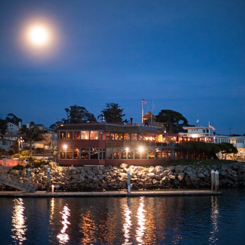 A waterfront restaurant illuminated at night, with a full moon above and reflections on the water.