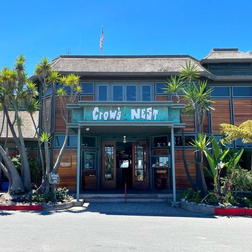 Exterior of the Crow's Nest restaurant, featuring wooden architecture and tropical plants under a clear blue sky.