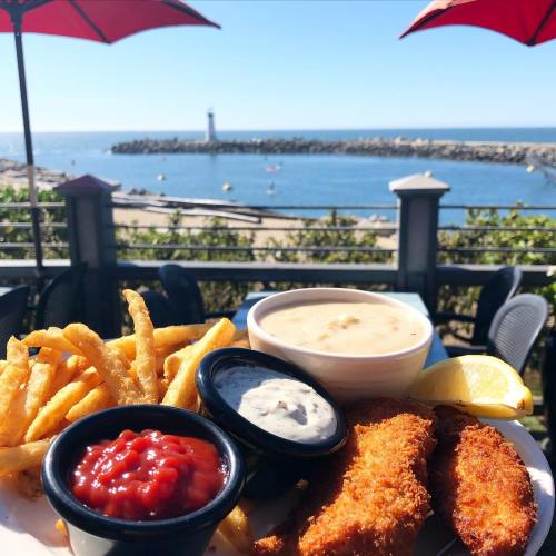 A plate of fried fish, fries, and dipping sauces with a scenic view of the ocean and a lighthouse in the background.