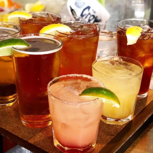 A variety of colorful cocktails and drinks with garnishes of lemon and lime on a wooden tray.