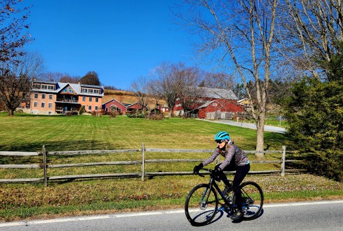 A cyclist in a blue helmet rides along a rural road with green fields and red barns in the background under a clear blue sky.