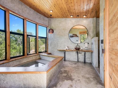 Modern bathroom with a wooden ceiling, large windows, a soaking tub, and a circular mirror. Natural light fills the space.