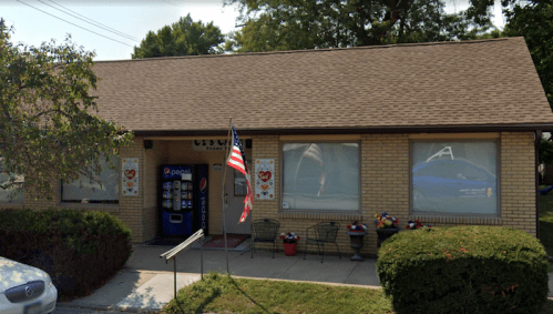 A brick building with a sloped roof, featuring windows, an American flag, and a vending machine outside.