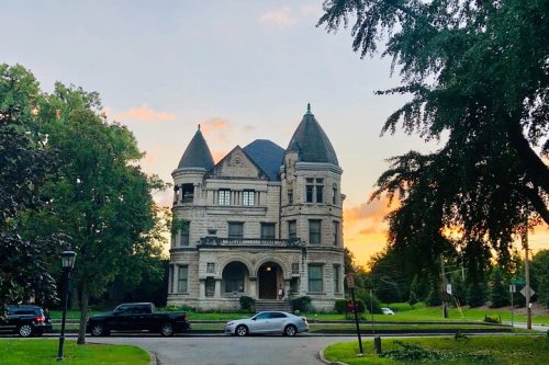 A large, historic mansion with turrets, surrounded by trees, at sunset.
