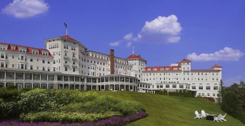 A grand white hotel with red roofs, set on a green hillside under a blue sky with fluffy clouds.