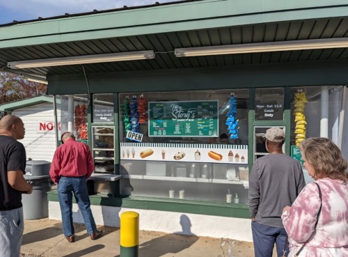 A food stand with a menu, colorful chip bags hanging, and customers waiting in line outside on a sunny day.