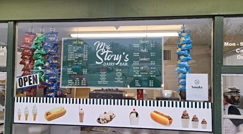 A storefront menu for Mrs. Story's Dairy Bar featuring hot dogs, shakes, and snacks, with colorful chip bags displayed.