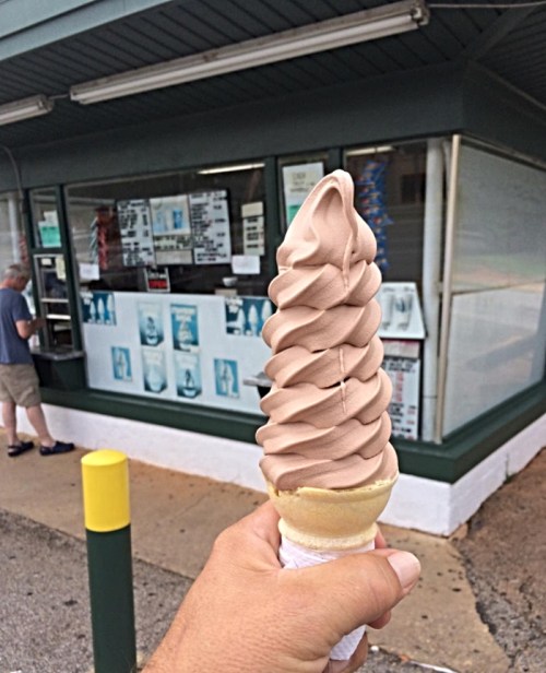 A hand holding a large chocolate soft serve ice cream cone in front of an ice cream shop.