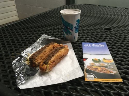 A hot dog wrapped in foil on a table, next to a drink and a brochure titled "100 Dishes to Eat in Alabama."
