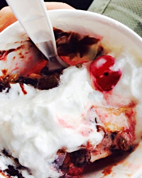 A close-up of a dessert cup filled with chocolate, whipped cream, cherries, and mixed fruits, with a spoon resting inside.
