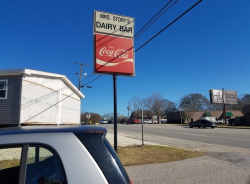 Sign for Mrs. Story's Dairy Bar with a Coca-Cola logo, alongside a road on a clear day. A car is parked nearby.