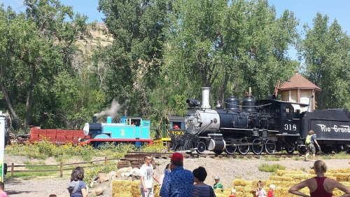 A blue cartoon train and a black steam locomotive are displayed in a park, with people enjoying the scene around them.