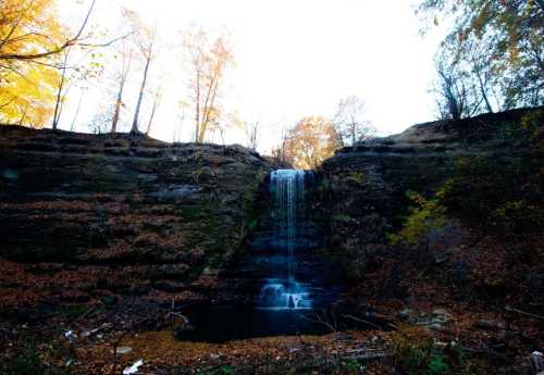 A serene waterfall cascading down rocky cliffs, surrounded by autumn foliage and a tranquil pool below.