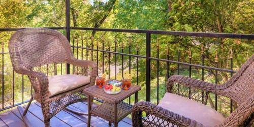 A cozy balcony with two wicker chairs and a small table, featuring drinks and snacks, surrounded by lush greenery.
