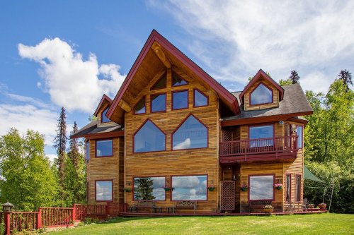 A large wooden house with a triangular roof, multiple windows, and a green lawn surrounded by trees under a blue sky.