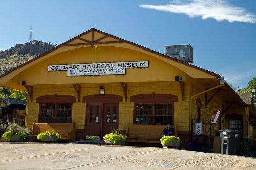 Exterior of the Colorado Railroad Museum, featuring a yellow building with a sign and benches outside.