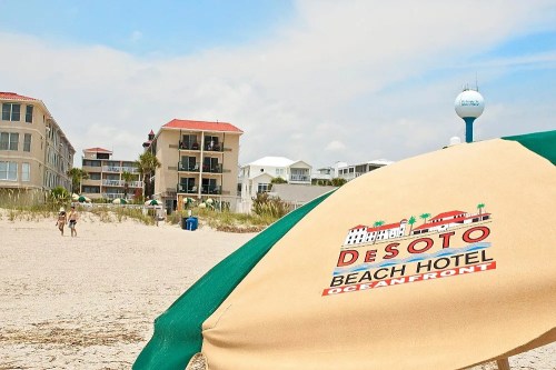 A beach scene featuring the Desoto Beach Hotel, with an umbrella in the foreground and people walking on the sand.
