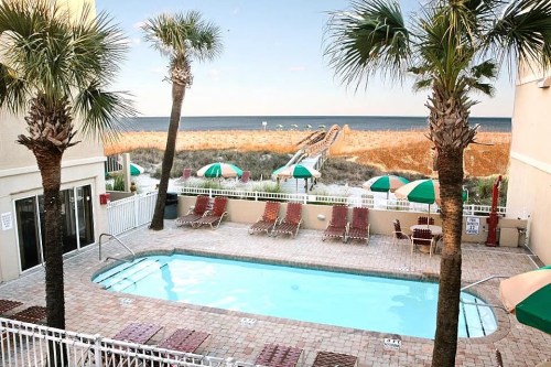 A pool area with lounge chairs and palm trees, overlooking a beach with umbrellas and a clear blue sky.