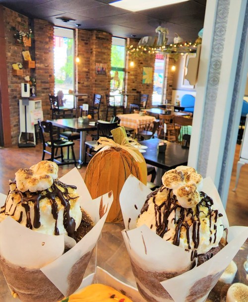 Two decadent desserts topped with whipped cream and chocolate drizzle, with a cozy café setting in the background.
