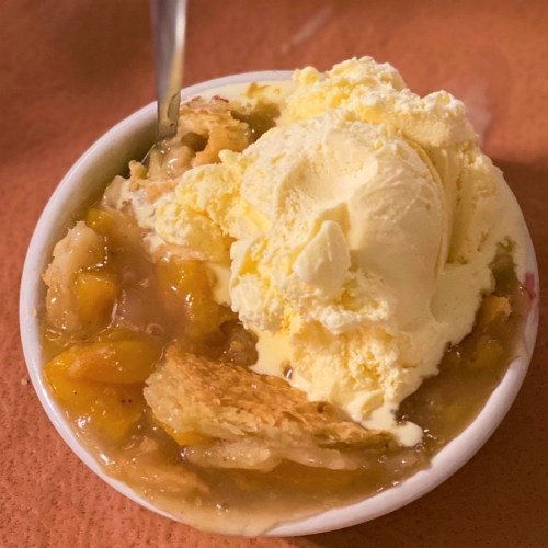 A bowl of peach cobbler topped with a scoop of vanilla ice cream and a spoon.