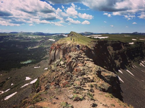 A rocky mountain ridge with hikers, surrounded by green valleys and distant peaks under a blue sky with clouds.