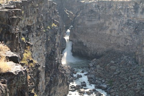 A narrow canyon with steep rocky walls and a waterfall cascading into a river below. Sunlight glimmers on the water.