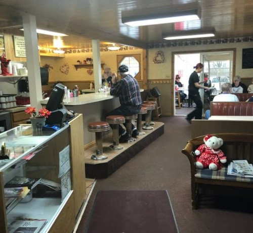A cozy diner interior with a counter, stools, and patrons enjoying their meals. A teddy bear sits on a nearby couch.
