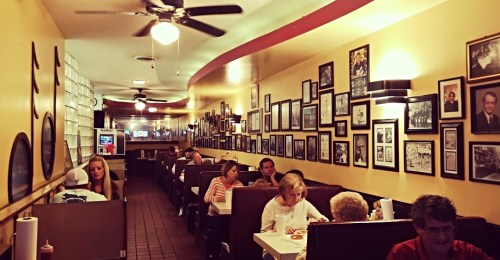 A cozy diner interior with patrons at booths, surrounded by framed photos and memorabilia on yellow walls.