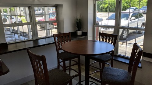 A small dining area with a round table and four chairs, featuring large windows overlooking parked cars outside.
