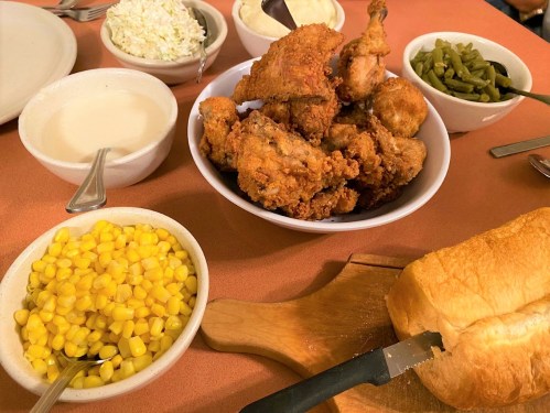A table spread with fried chicken, corn, green beans, coleslaw, gravy, and sliced bread on a cutting board.