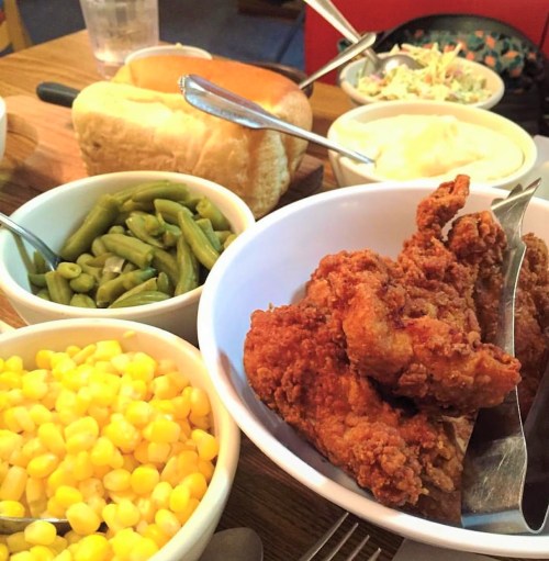 A table spread featuring fried chicken, green beans, corn, mashed potatoes, coleslaw, and bread rolls.