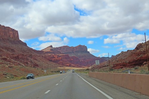 A scenic highway stretches through red rock formations under a partly cloudy sky.