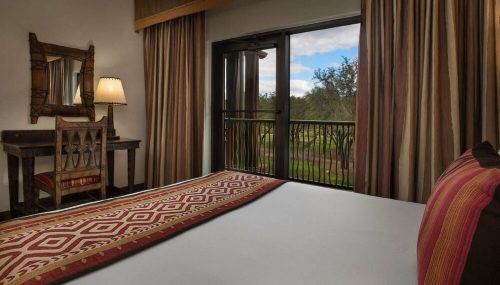 Cozy hotel room with a patterned bedspread, a wooden desk, and a view of greenery through large glass doors.