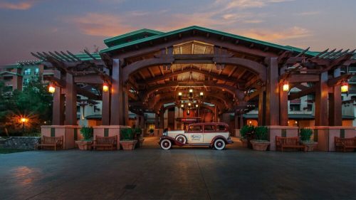 A vintage car parked under a wooden archway at a resort entrance, illuminated by warm lights at sunset.